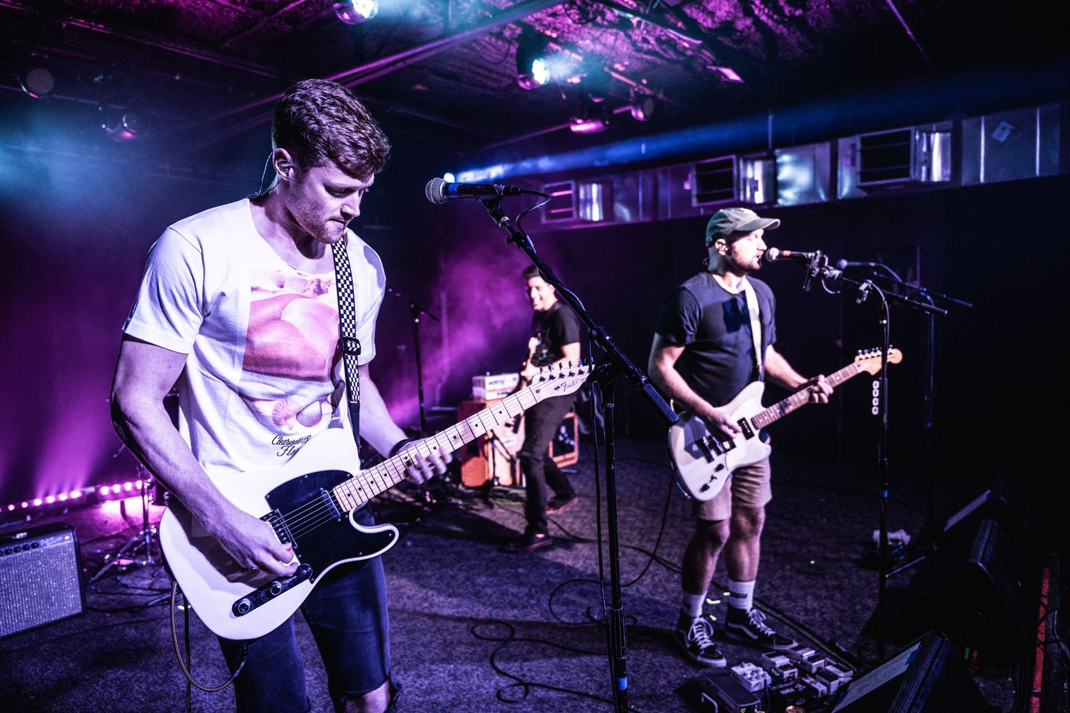 Rock band performs energetically on stage with guitarists under purple and blue lighting at small venue.