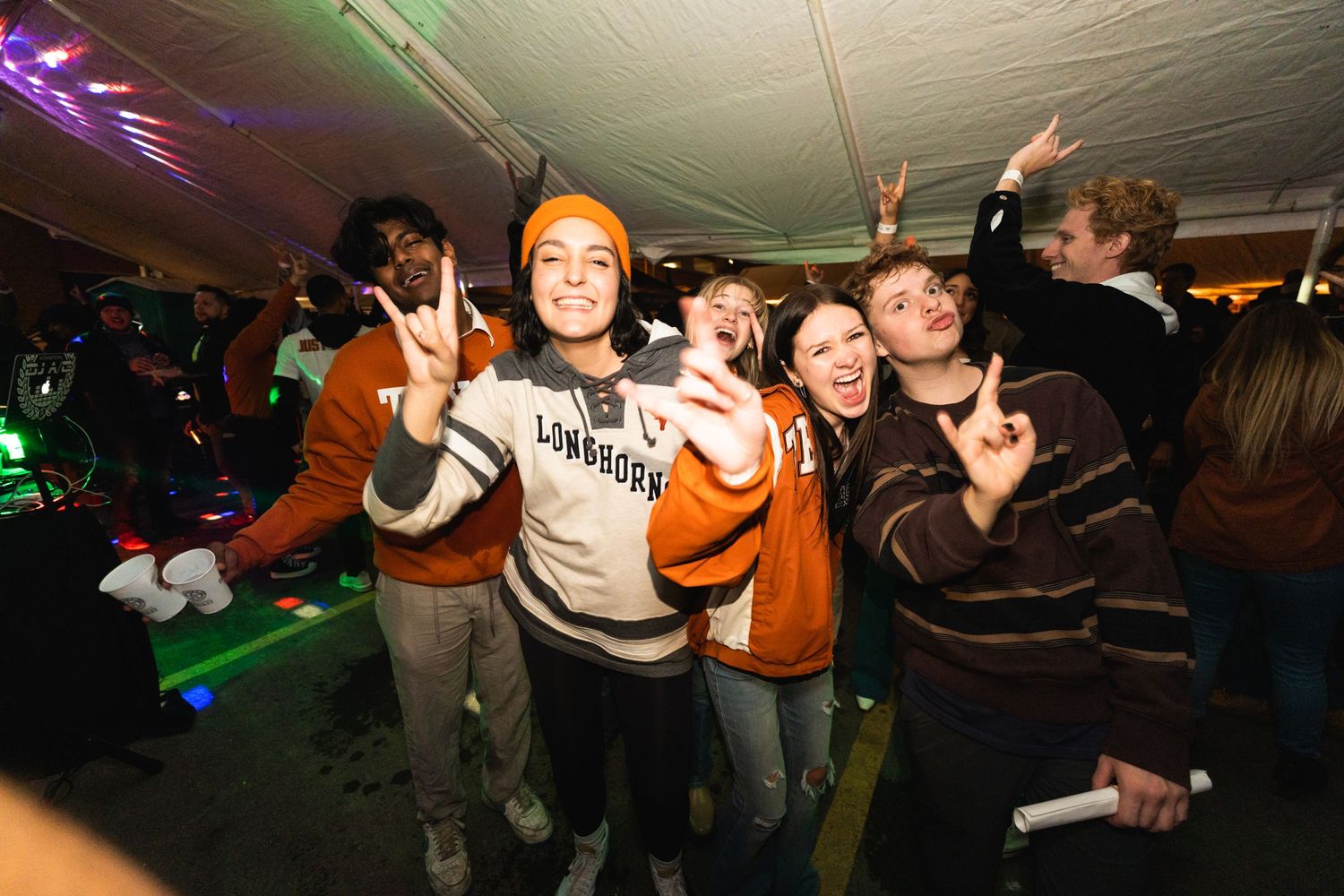 A group of energetic party-goers dancing and celebrating under colorful lighting at a nighttime event.