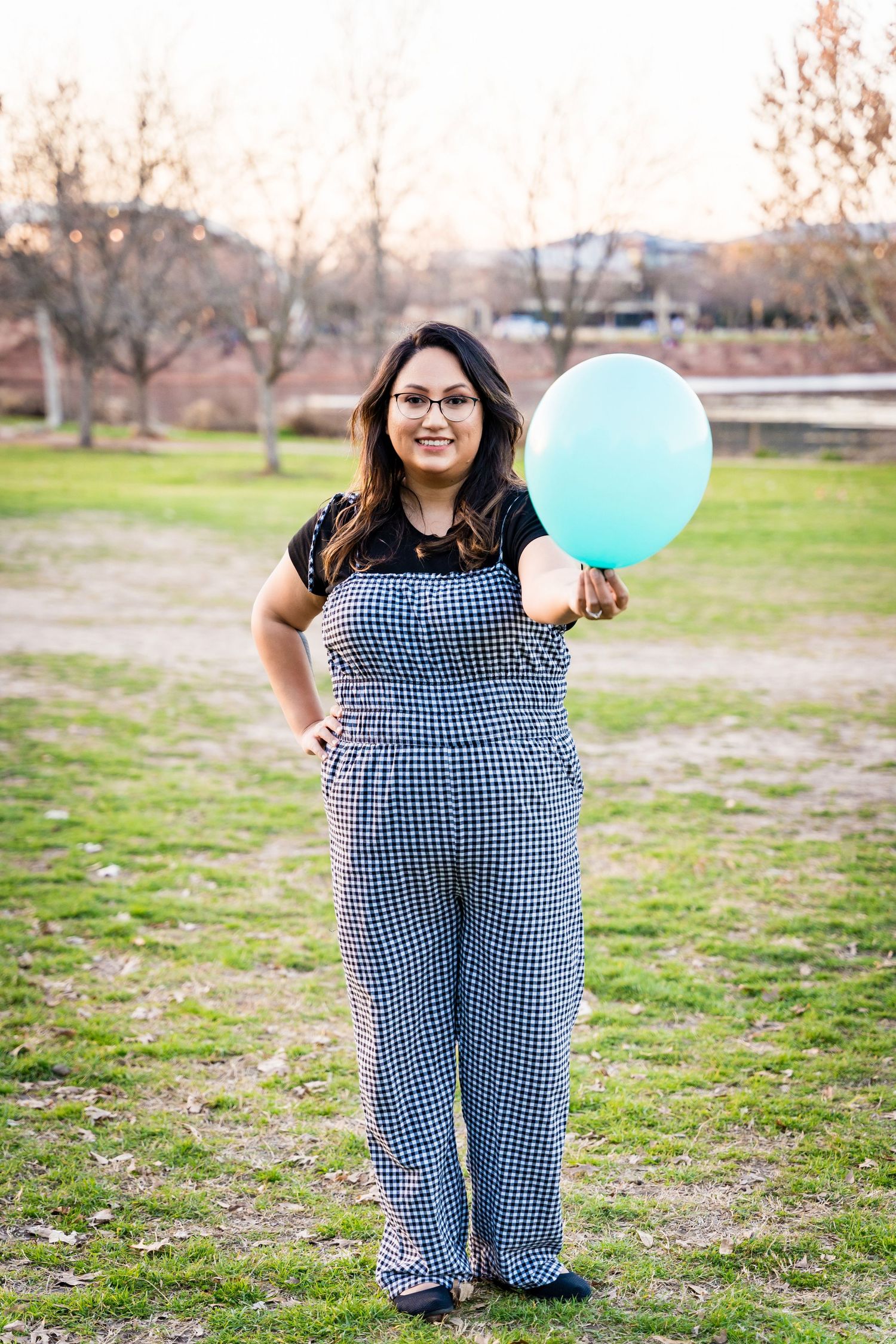 Person in gingham overalls holding a mint green balloon in a park setting at sunset.