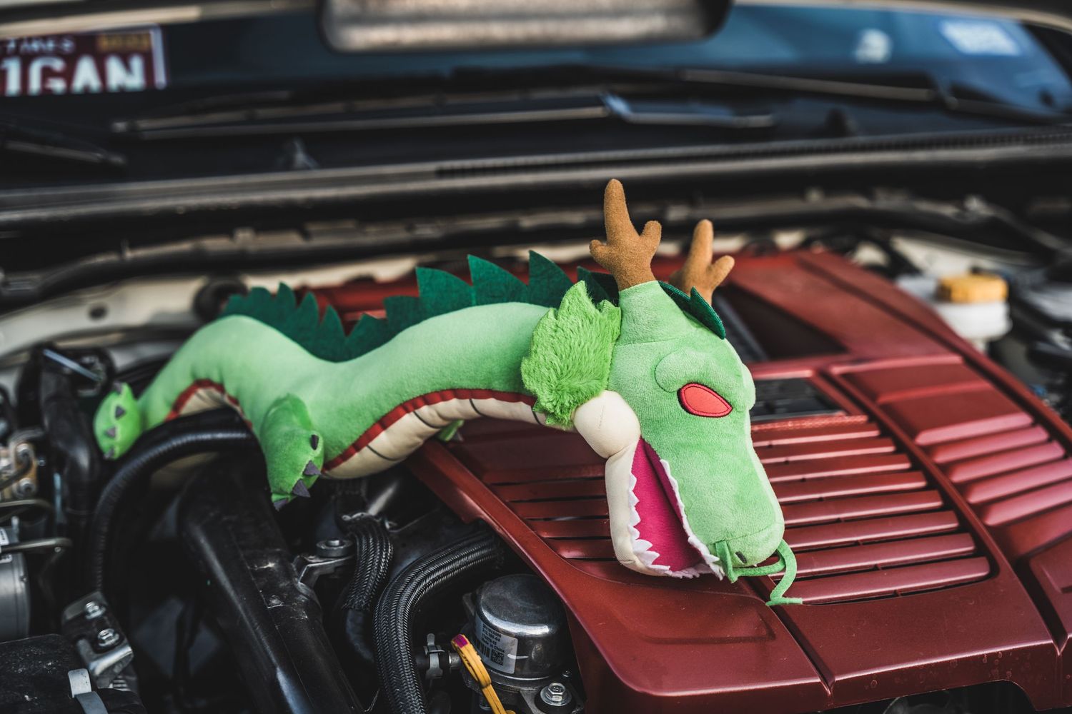 A playful green dragon plush toy resting on a car engine bay.