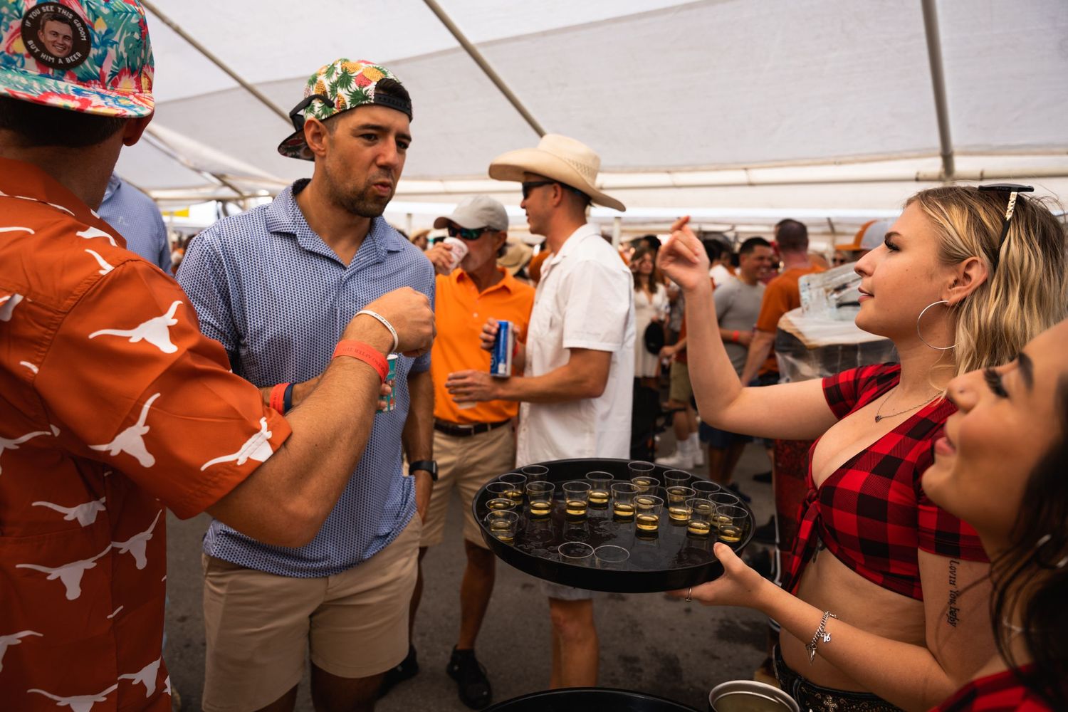 Fans enjoying drinks and conversation under a white tent at a tailgate party.