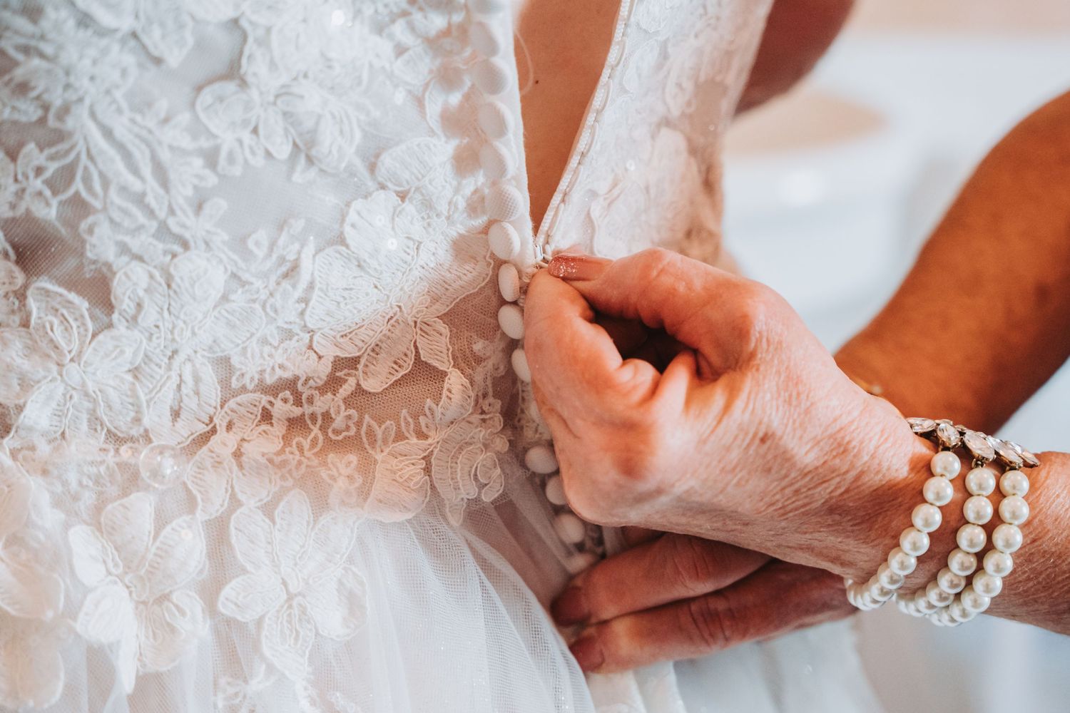 Close-up of hands fastening buttons on a detailed white lace wedding dress with pearl bracelets.