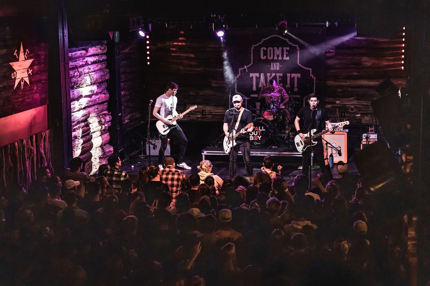 Band performs on stage with rustic wooden walls and enthusiastic crowd below.