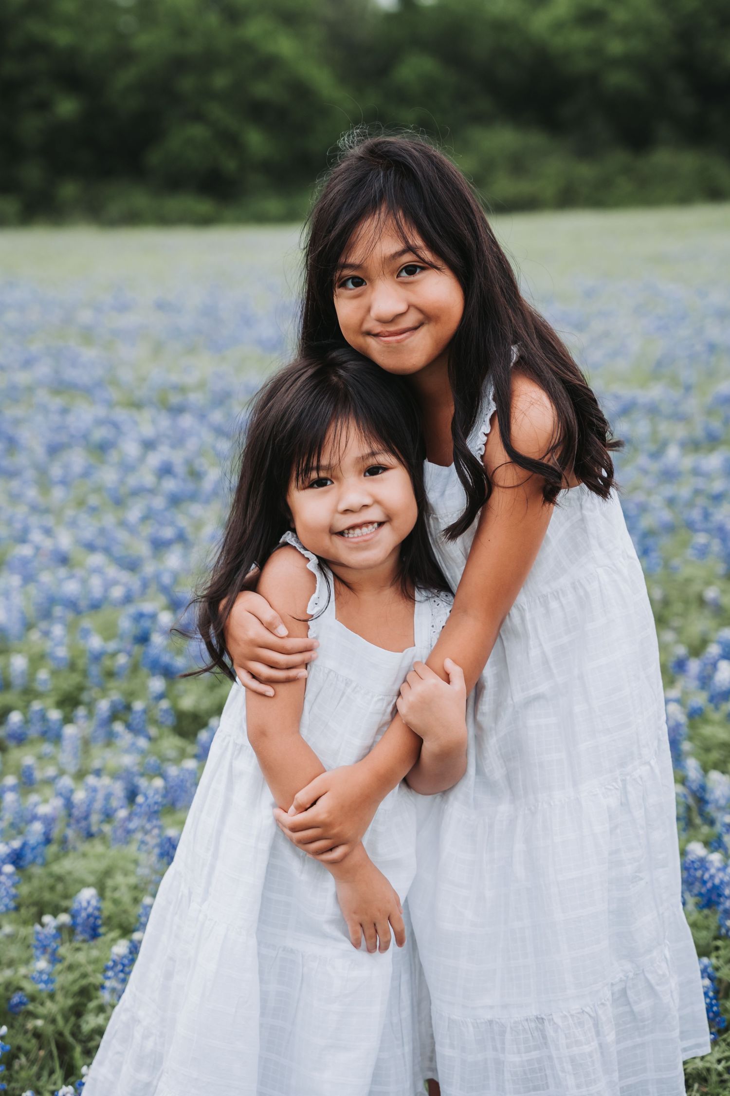 Two figures in white dresses embrace while standing in a field of Texas bluebonnets.