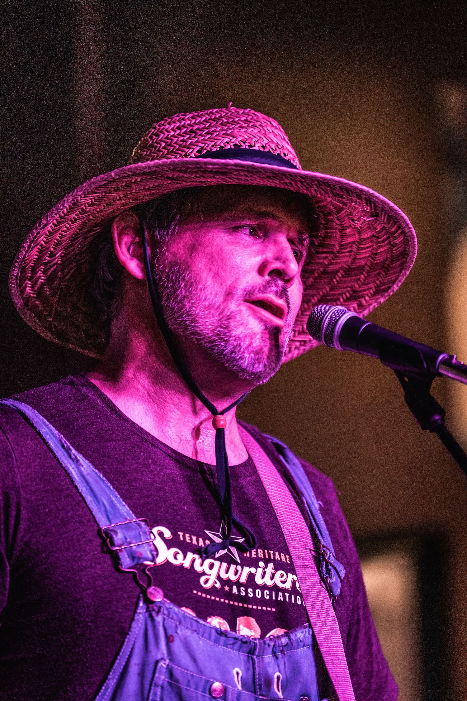Musician wearing straw hat and overalls performs vocals under purple stage lighting.