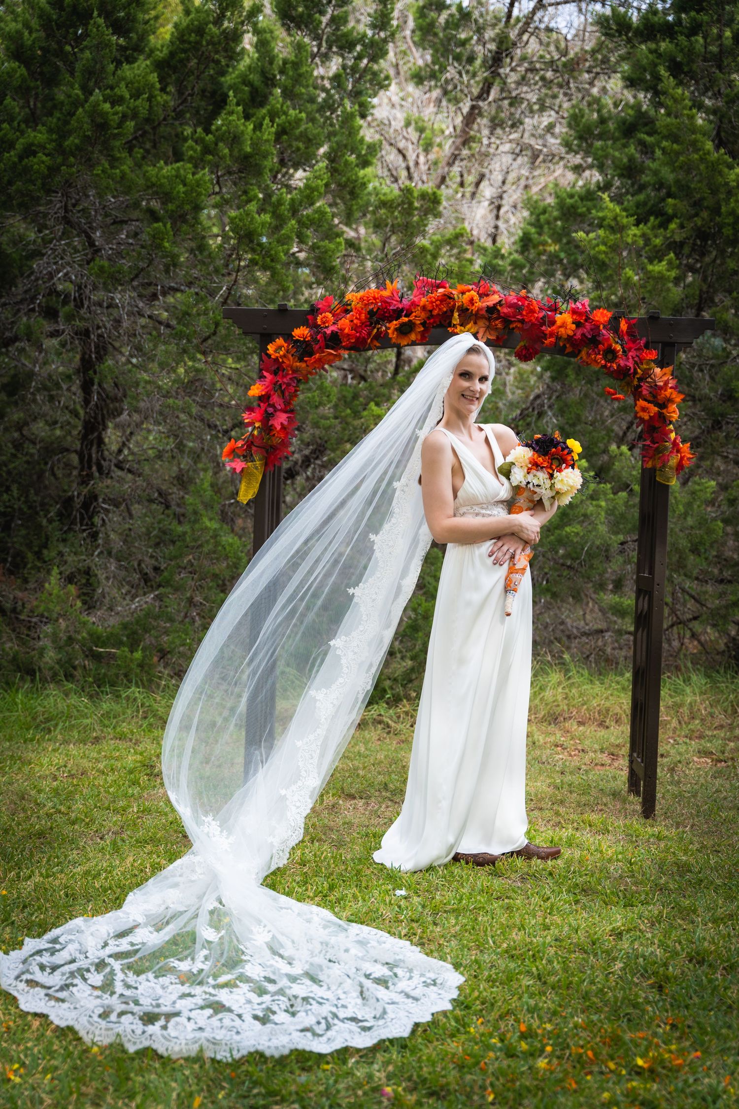 A bride in a flowing white dress and veil poses under a vibrant red floral arch in an outdoor garden setting.