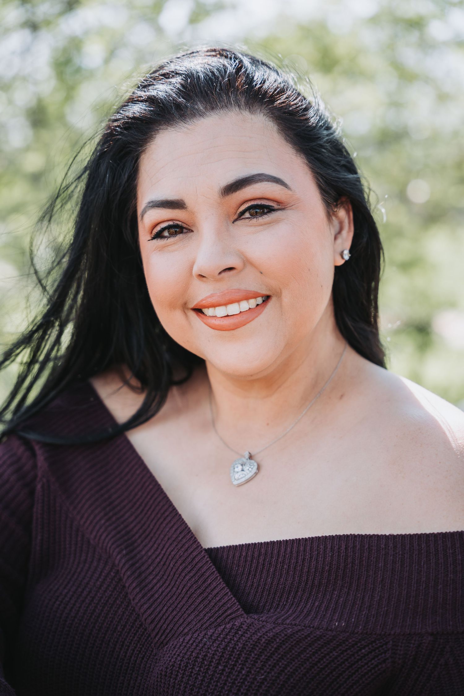 Person in burgundy sweater smiles warmly in outdoor portrait with bokeh background.