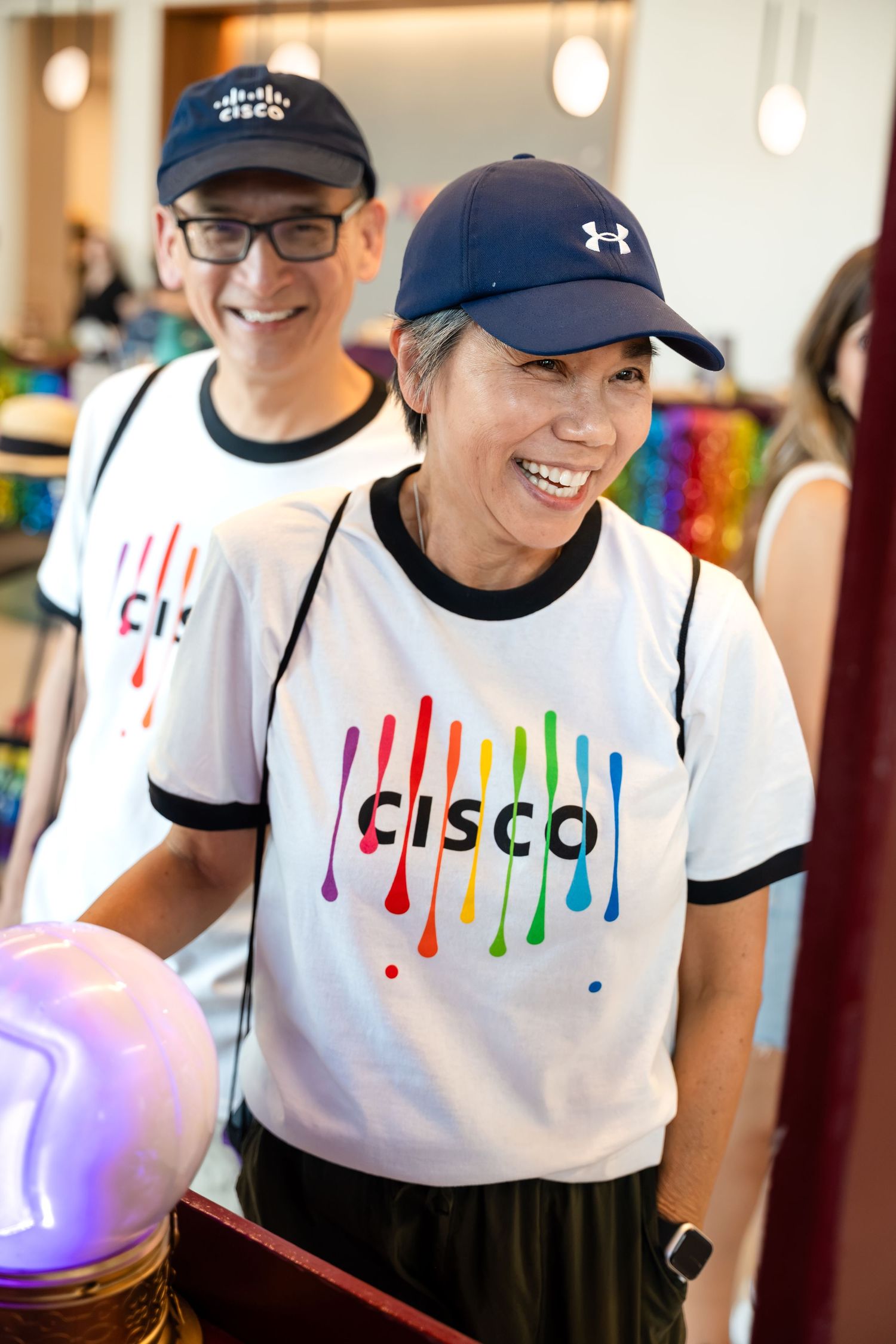 Two smiling Cisco employees wearing rainbow pride logo t-shirts and caps at a colorful company event.