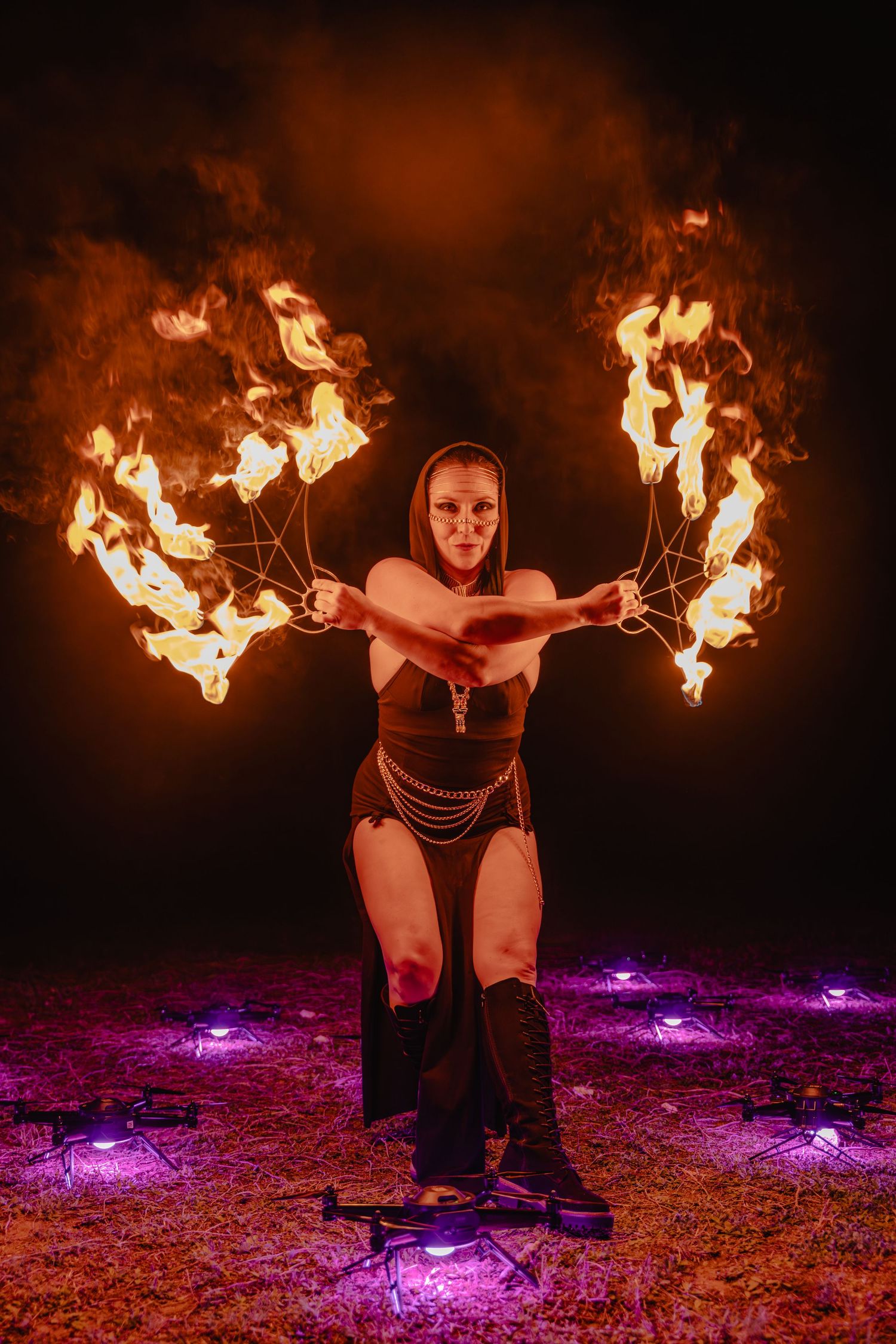 Fire dancer performs with flaming fans surrounded by drones and purple lights at night outdoor show.