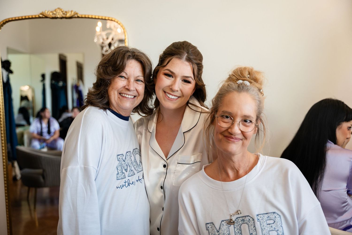 Three women smiling together, two wearing matching MOH shirts, celebrating a bridal party getting ready.