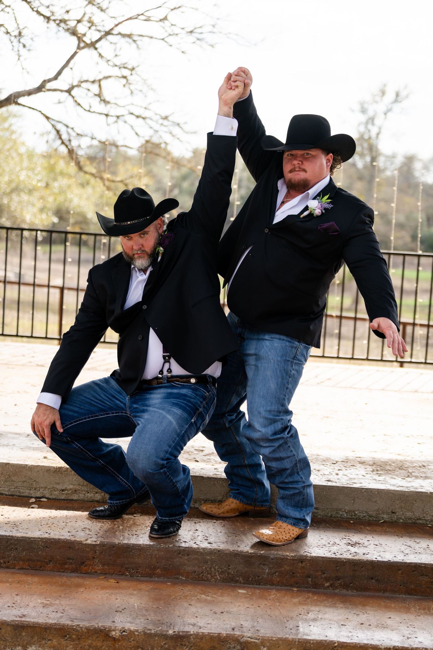 Two grooms in black blazers, cowboy hats, and jeans pose playfully outdoors at a country wedding.