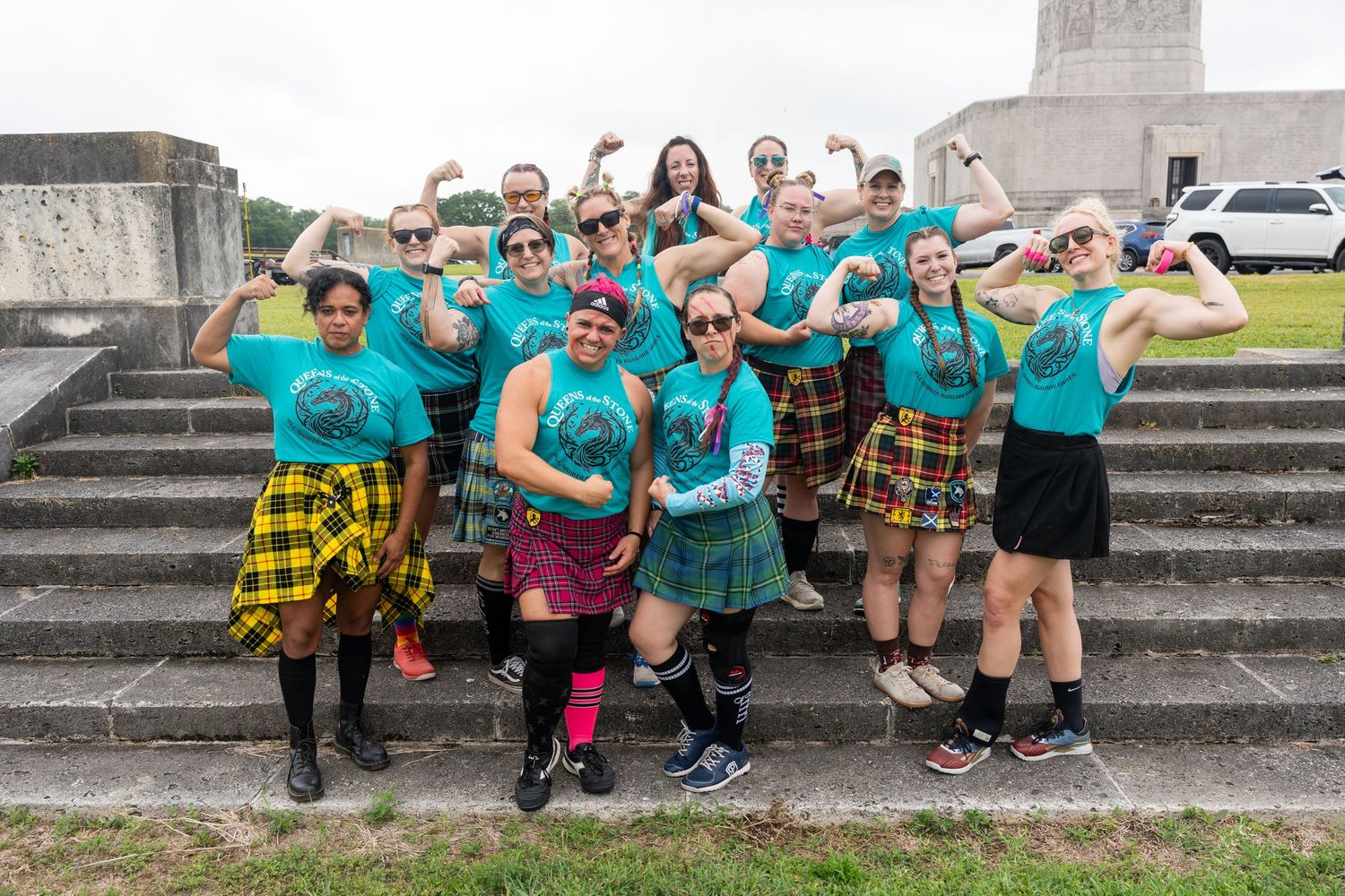 Group of women in teal shirts and kilts flexing muscles on stone steps at outdoor event.