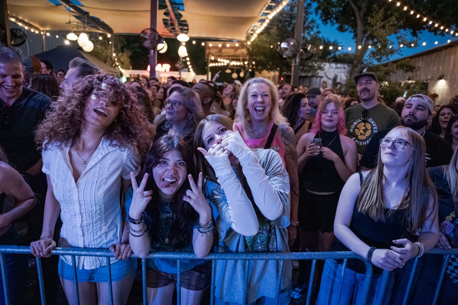 Excited crowd of fans at an outdoor music festival at night, cheering behind a barrier.
