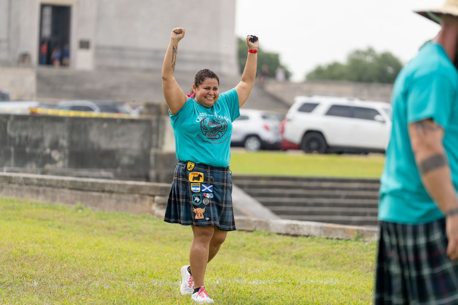 Woman in teal shirt and tartan kilt celebrating with arms raised at a Highland Games competition outdoors.