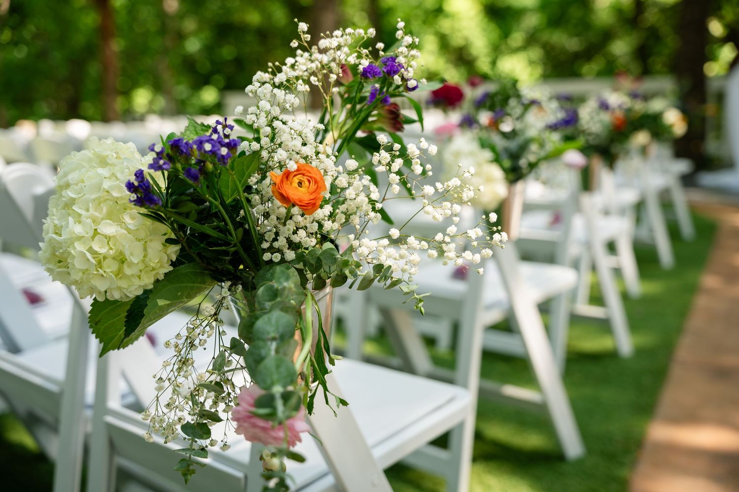 Elegant outdoor wedding aisle with white chairs adorned with colorful floral arrangements featuring hydrangeas and ranunculus.