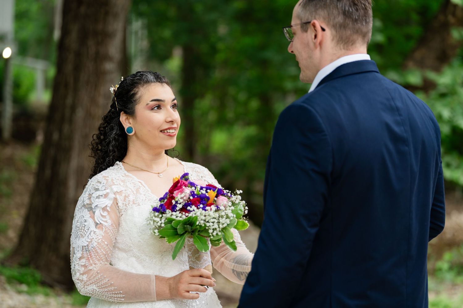 Bride in lace gown holds colorful bouquet, smiling at groom in navy suit during outdoor wedding ceremony.