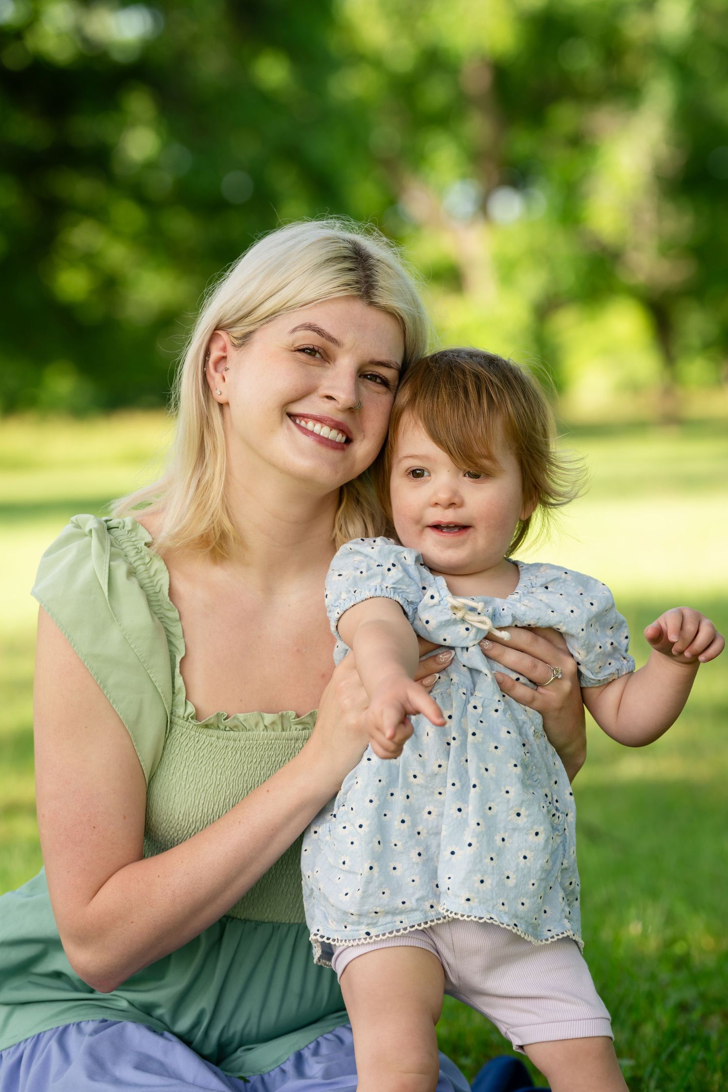 Smiling blonde mother in green dress holding toddler girl in white polka dot dress in sunny park.