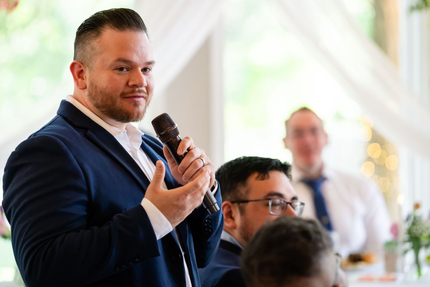 Man in navy suit giving a speech at a wedding reception while holding a microphone.