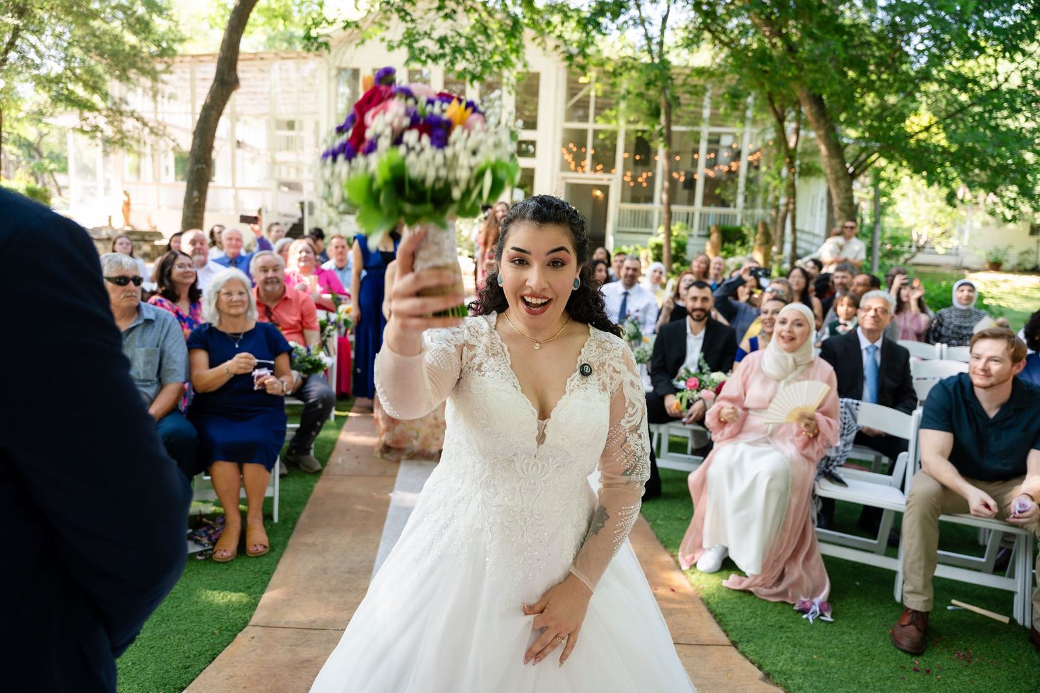 Bride in white gown tossing colorful bouquet to guests at outdoor wedding ceremony.