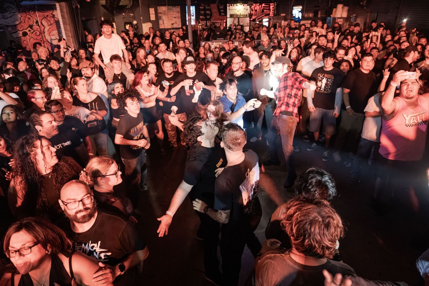 Large crowd of people dancing and socializing at a lively indoor concert or bar event with dim lighting.