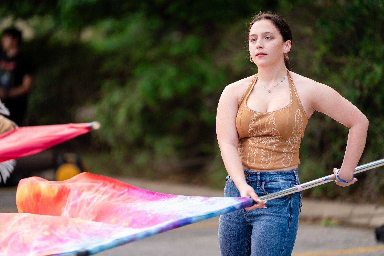Young woman in brown halter top and jeans performing color guard with colorful flag at outdoor parade.