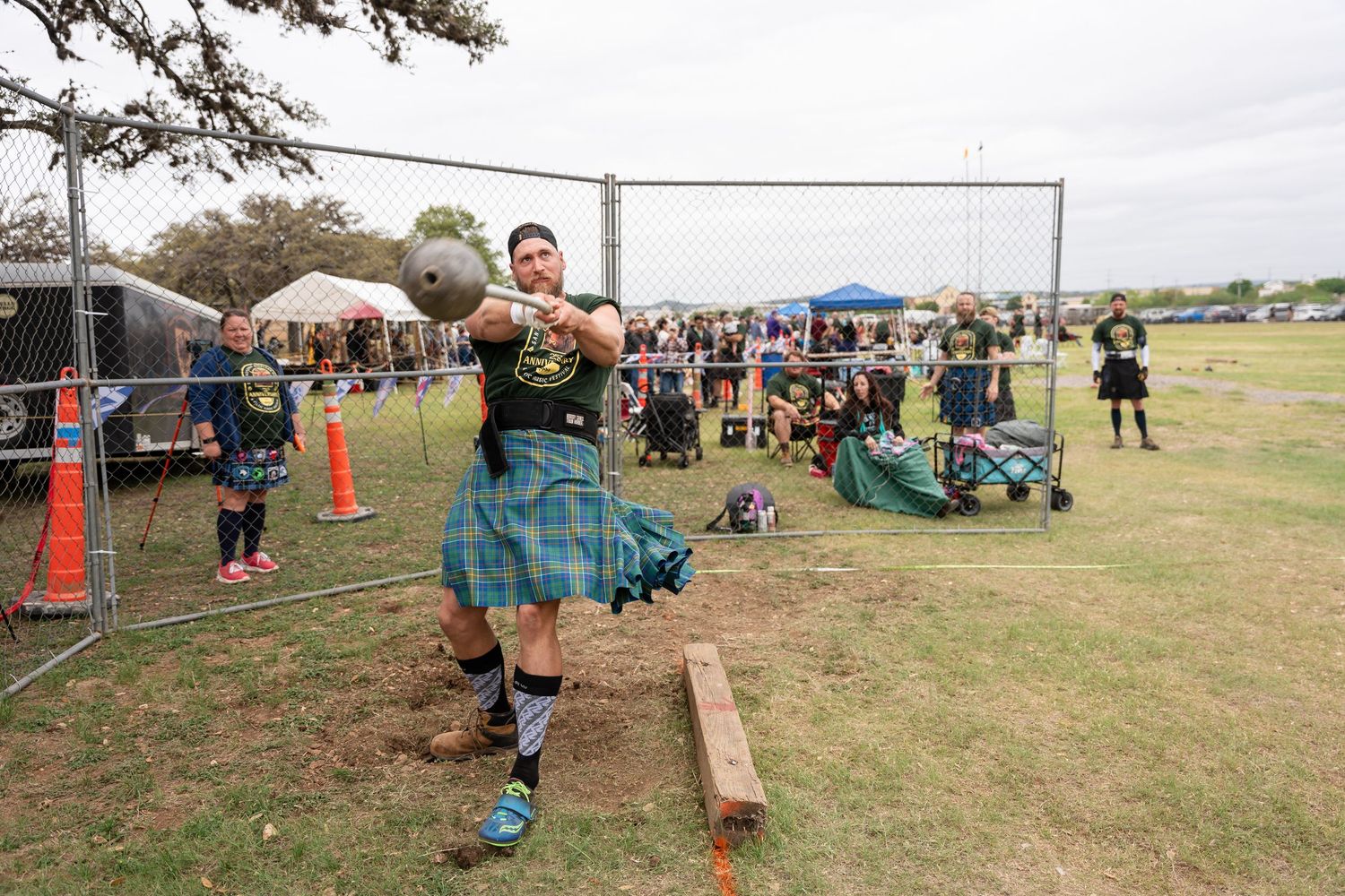 Man in blue tartan kilt throwing a weight at a Highland Games competition outdoors.