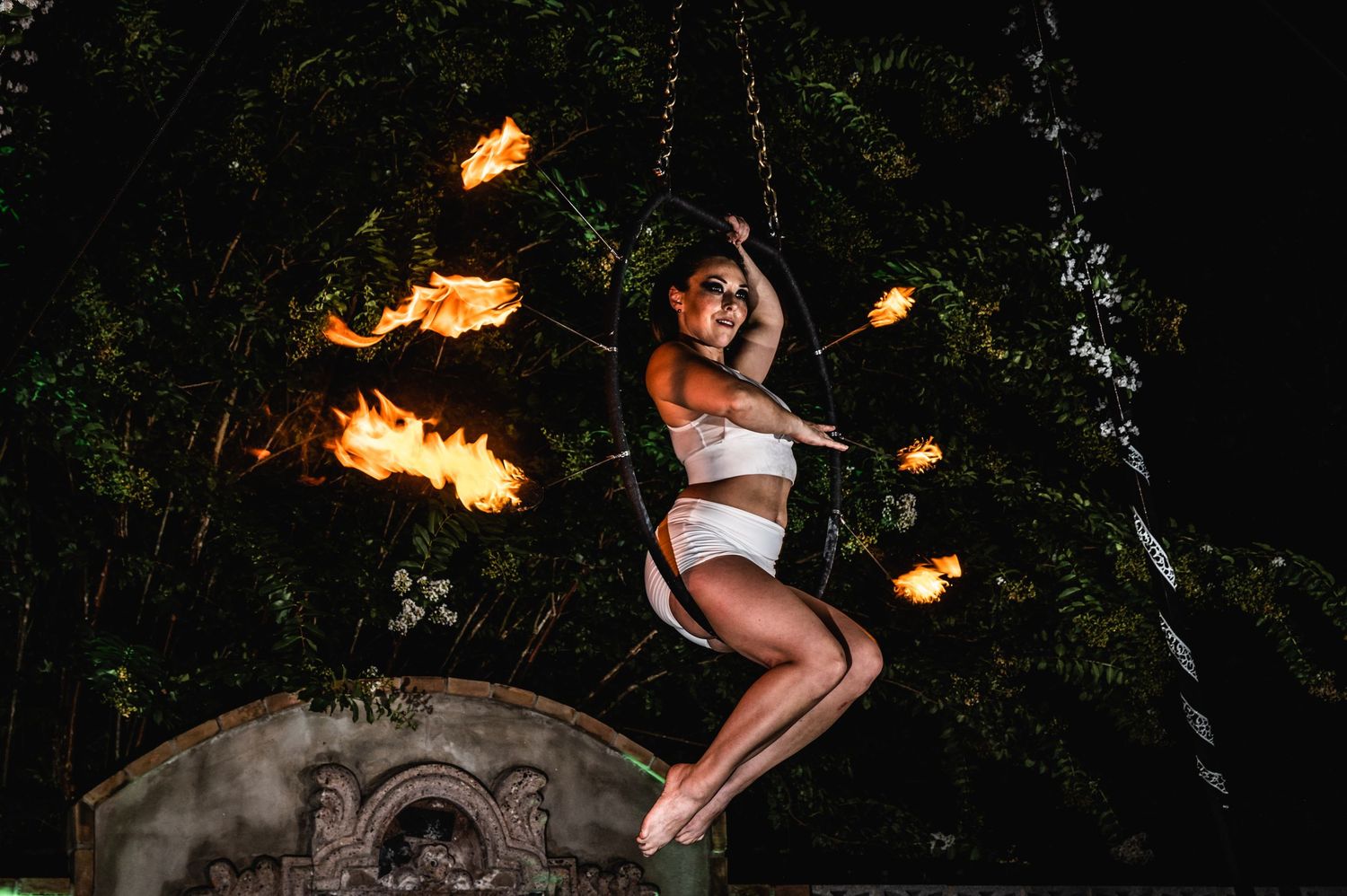 A performer in white outfit swings through the air surrounded by dramatic fire trails in a dark forest setting.