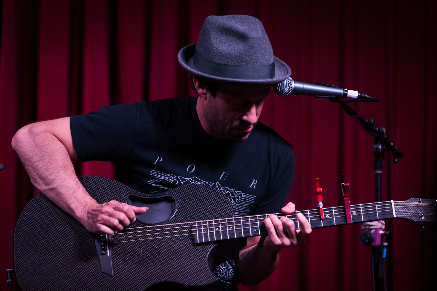Close-up view of a guitarist performing on stage with a dark hat and acoustic guitar in red lighting.