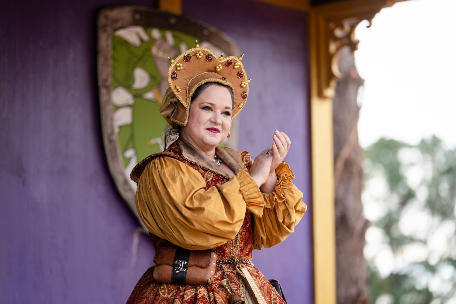 A person in a golden Renaissance-style dress and headdress poses elegantly against a purple wall in a historical setting.