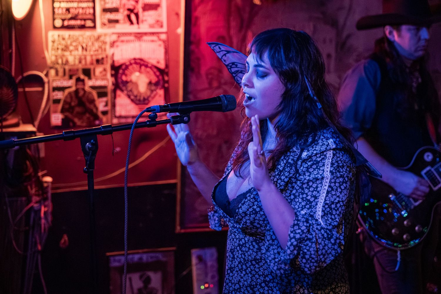A performer sings passionately into a microphone on stage bathed in dramatic red and purple lighting at a music venue.