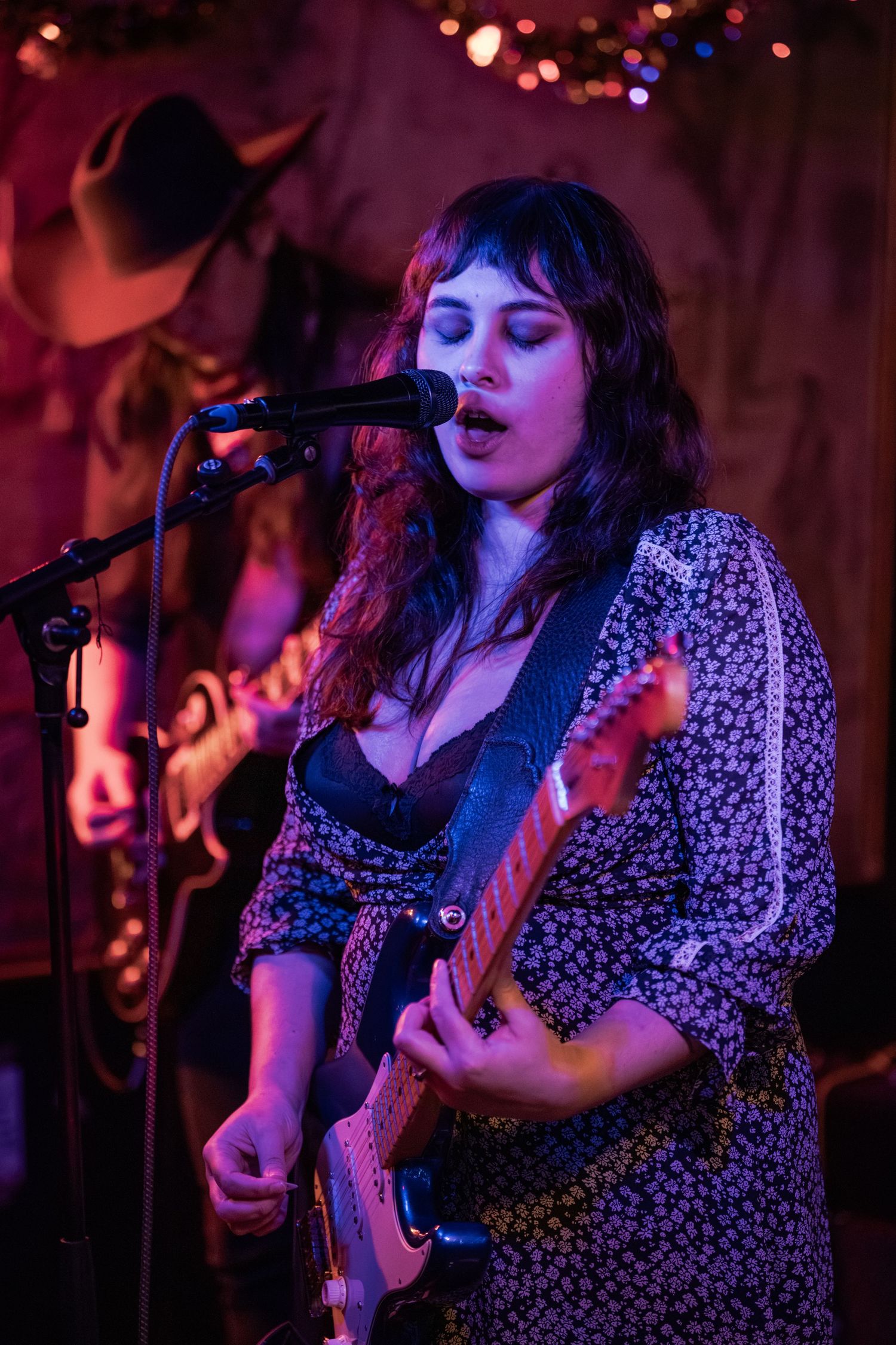 A musician performs on stage in a floral print dress with a guitar under moody purple lighting at a live music venue.