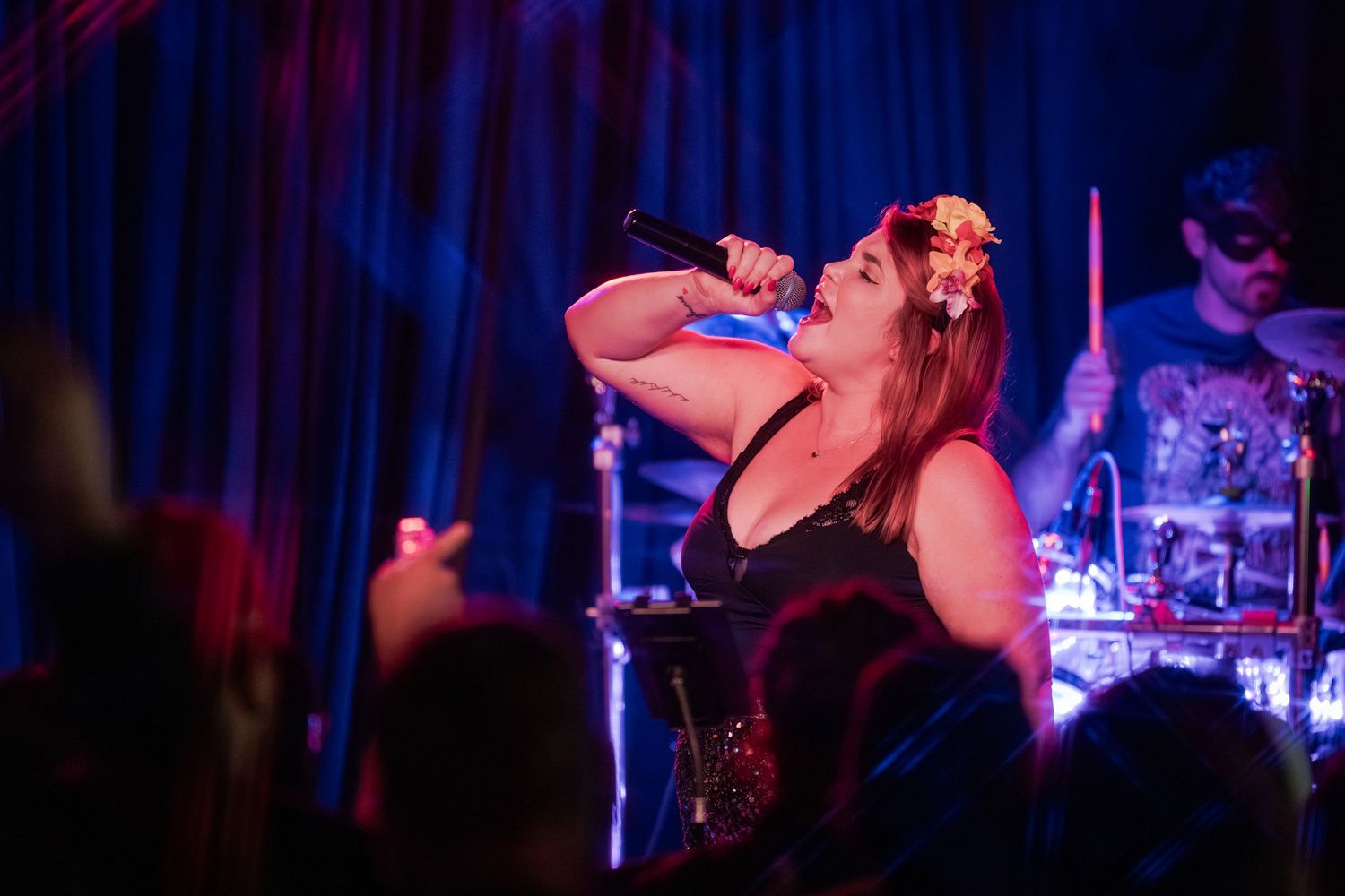 A performer in a dark venue sings passionately into a microphone with blue and purple stage lighting in the background.