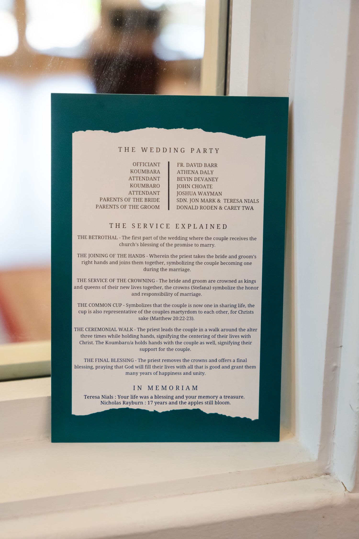 Wedding ceremony program details printed on white paper with dark teal border displayed in window light.
