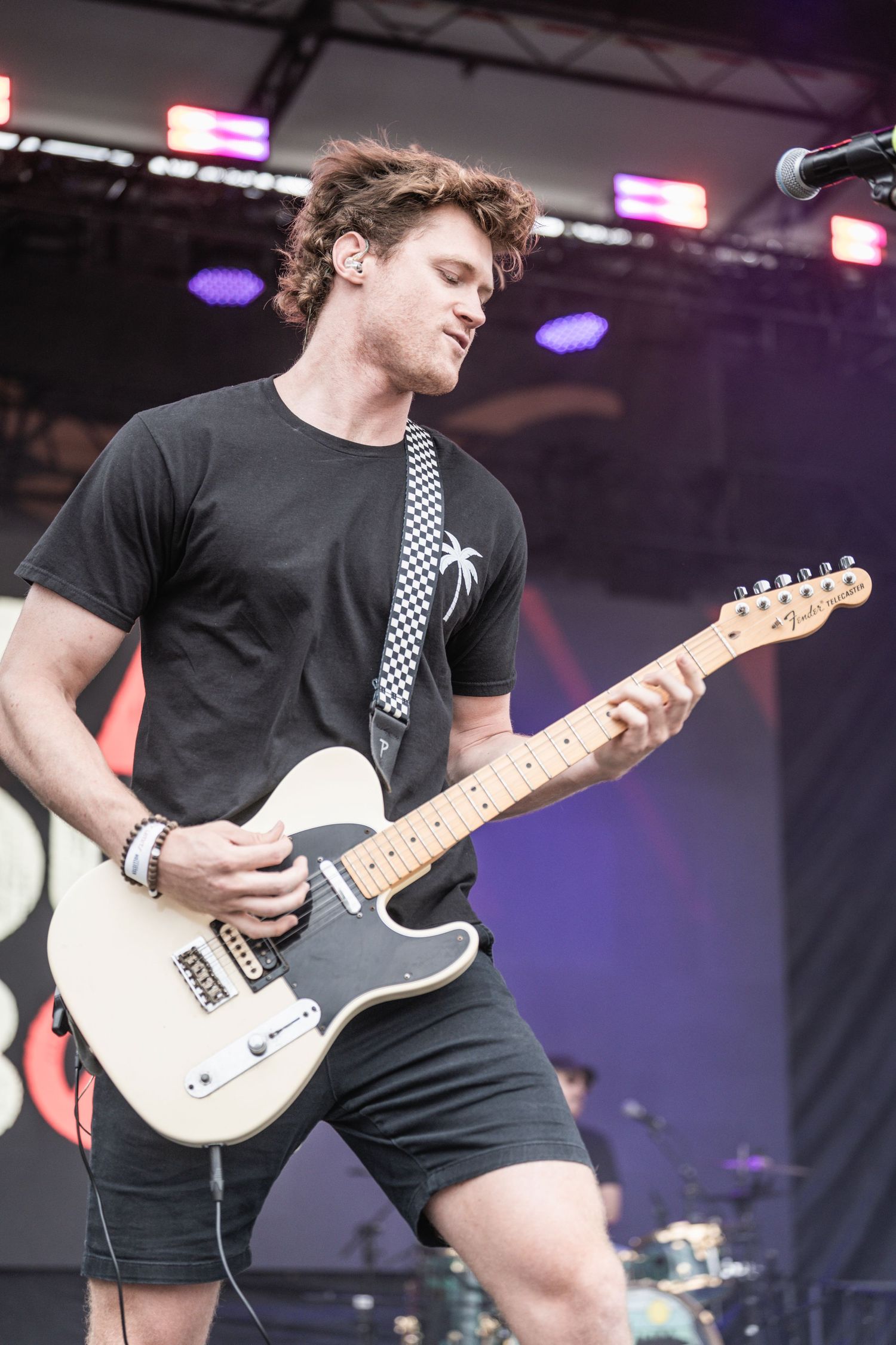 Guitarist in black t-shirt with checkered strap performs energetically on stage under purple lighting.