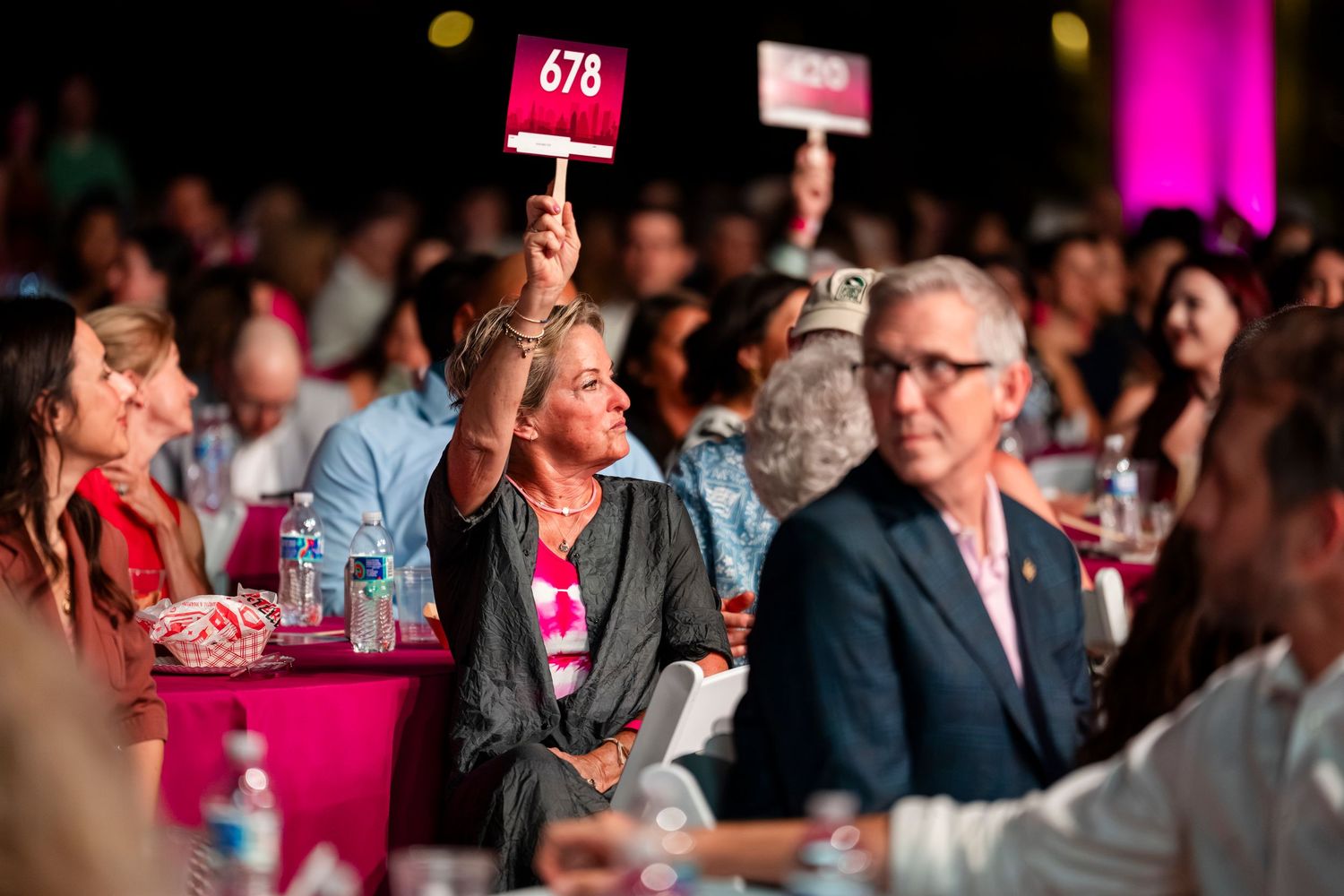 Audience members hold up numbered bidding paddles during a crowded indoor auction event with pink lighting.