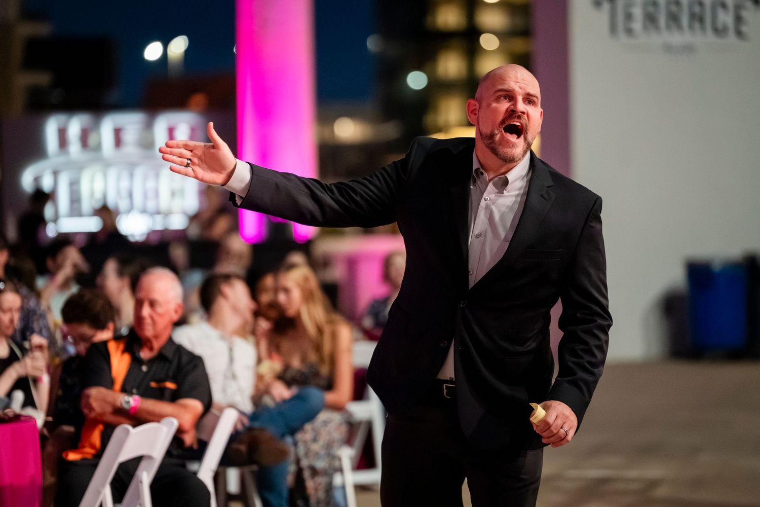 A person in a suit gestures enthusiastically while speaking on stage at a business event with colorful lighting.