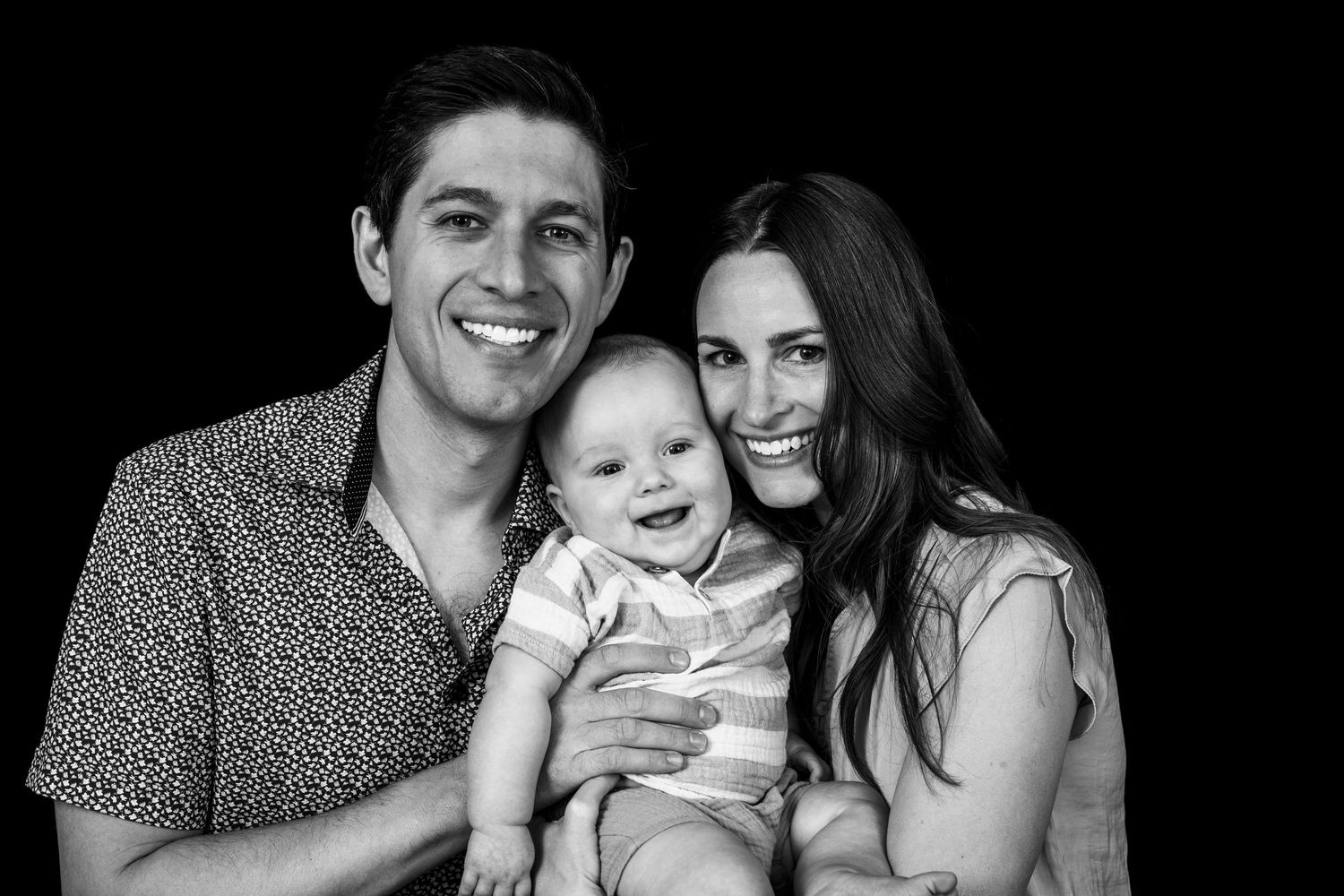 A black and white portrait of a couple holding their smiling baby between them.