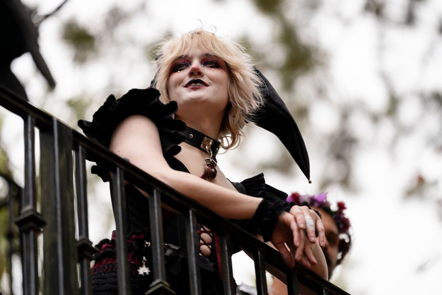 A person with blonde hair poses dramatically against a blurred outdoor background in black attire.