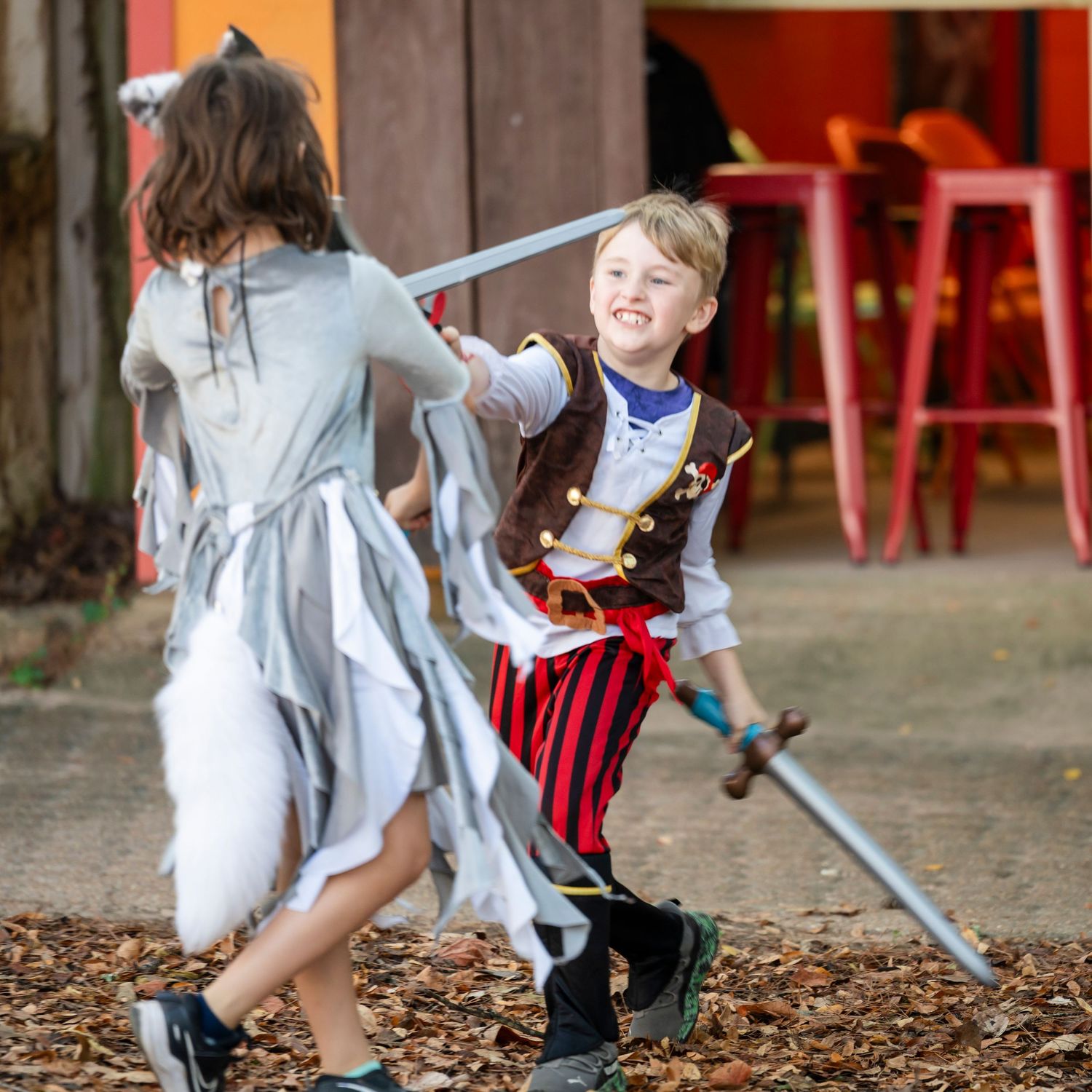 Two children play sword fighting with toy swords while wearing costumes on a ground covered with autumn leaves.
