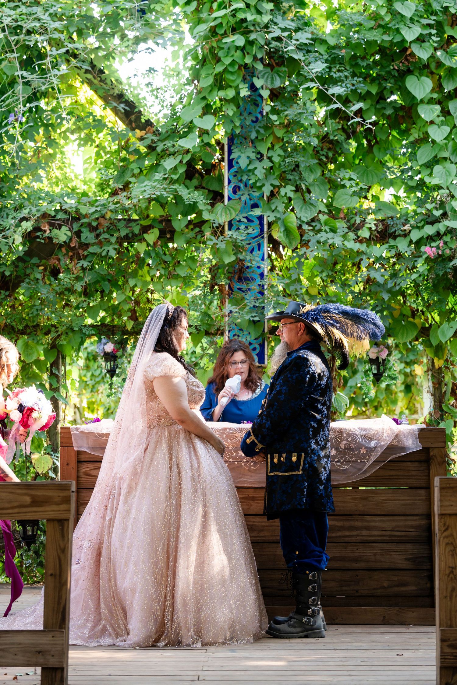 A couple in vintage-inspired attire exchanges vows during an outdoor garden ceremony on a wooden deck.