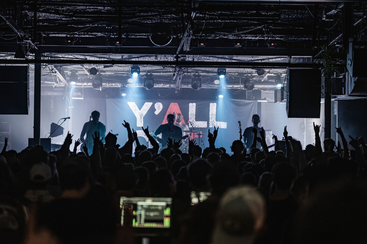 Silhouetted crowd at a concert with illuminated Y'ALL sign and blue stage lighting creating an energetic atmosphere.