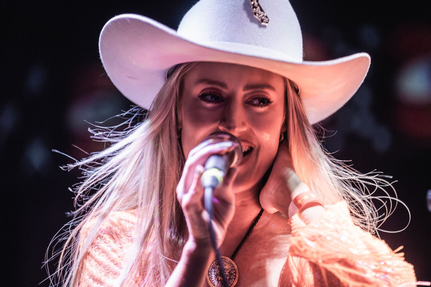 Country music performer in white cowboy hat and pink outfit sings into microphone during stage performance.