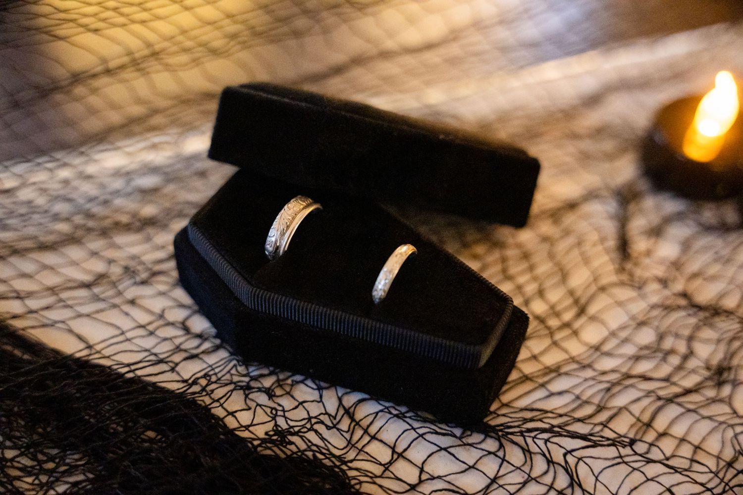 Two wedding rings displayed in a black coffin-shaped box on a textured surface with candlelight.