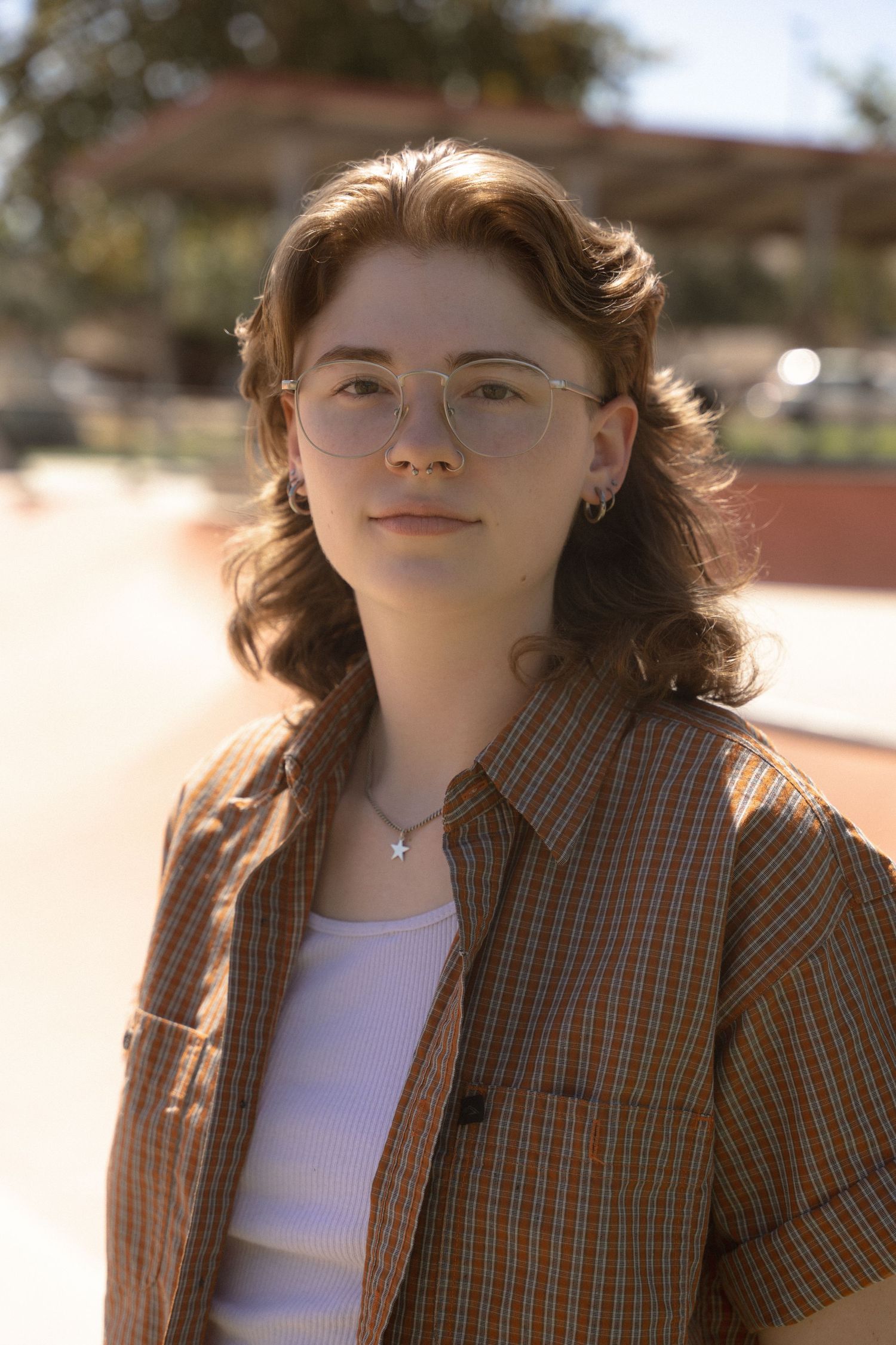 A person in a plaid button-up shirt and glasses poses outdoors in warm sunlight.