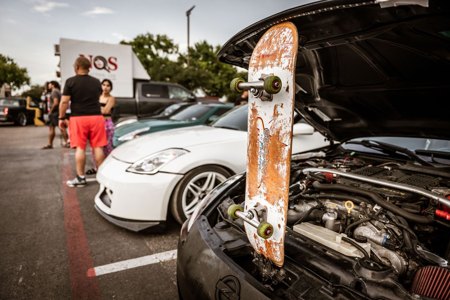 Used skateboard leaning against car with white sports car in background at automotive meet.