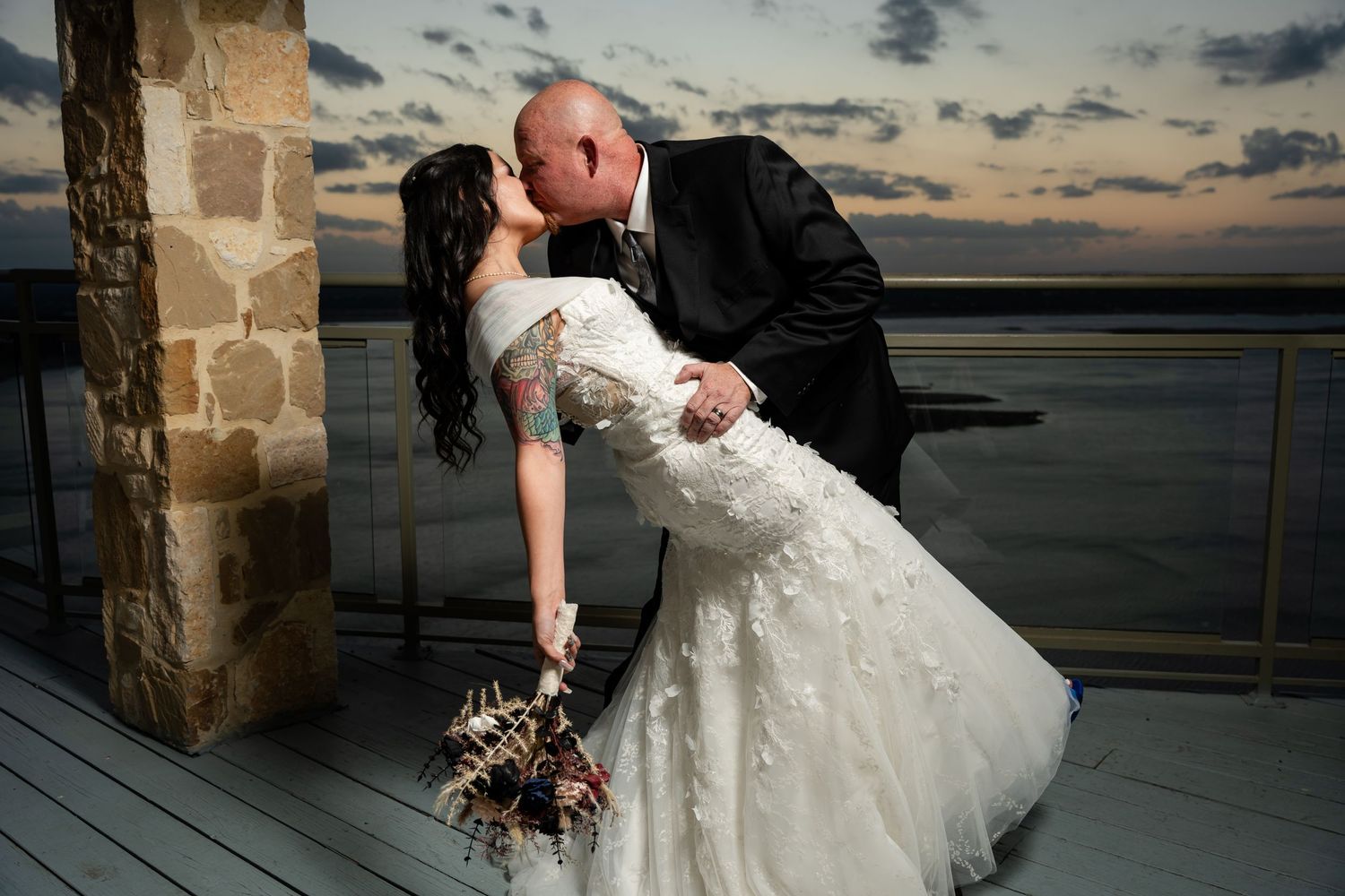 Romantic sunset wedding photo on a waterfront deck with stone architecture.