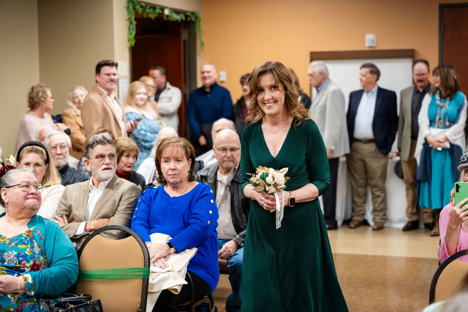 Guests gather for a celebratory event in a community room with someone in an elegant dark green dress at the center.