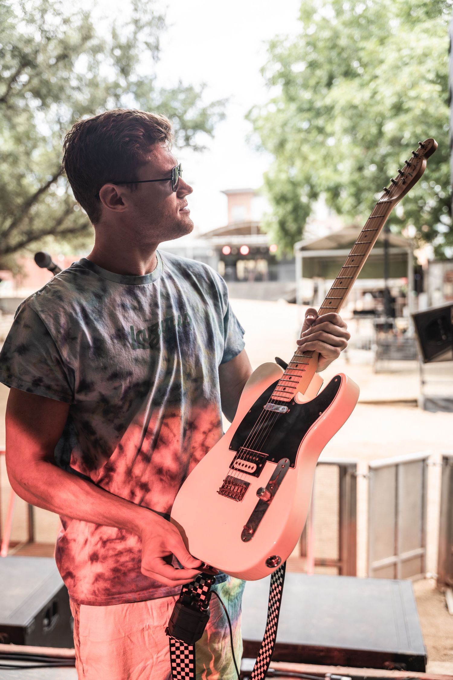 Musician in tie-dye shirt holding a pink electric guitar outdoors at a venue.