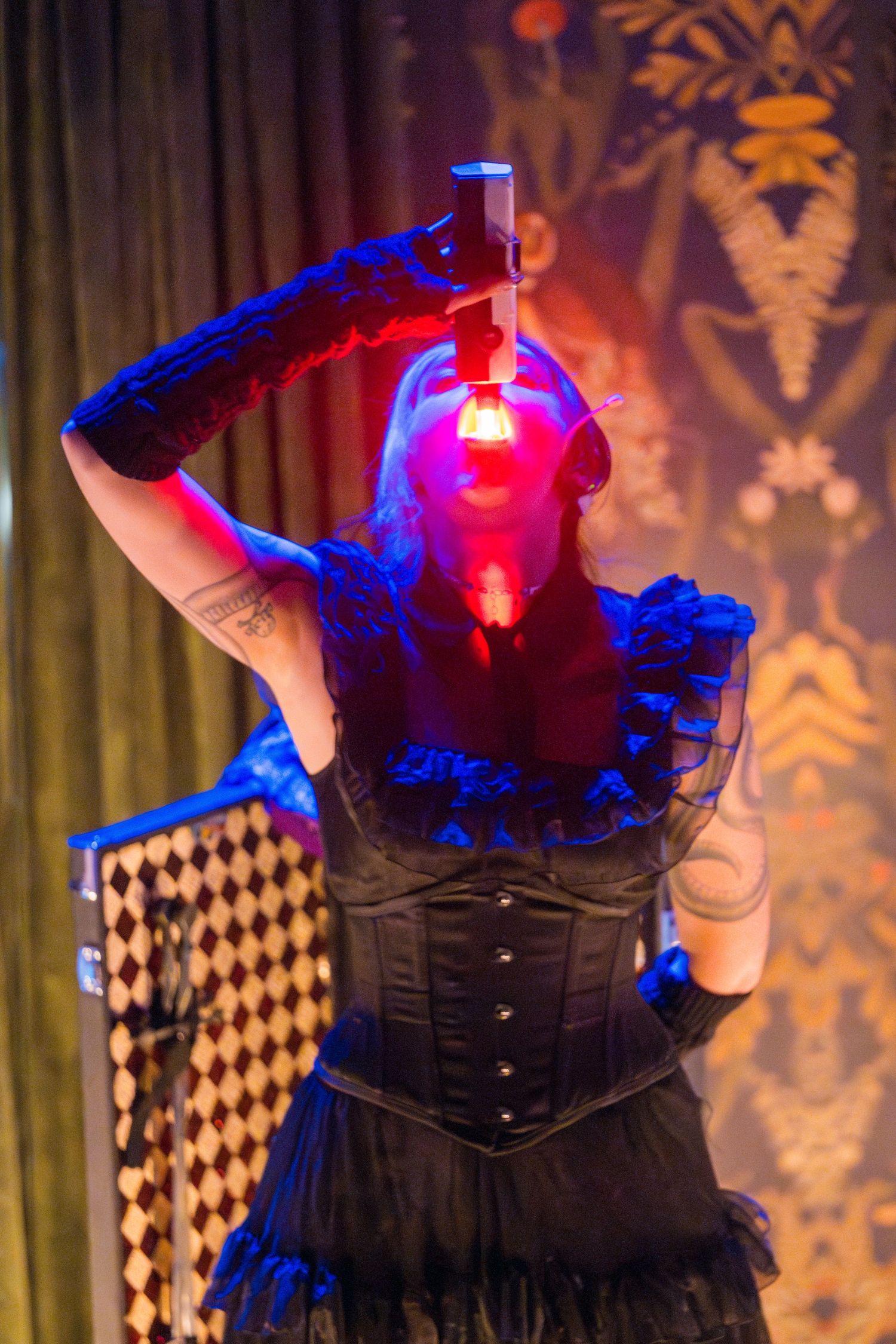 A performer in a black corset costume poses dramatically under red and blue stage lighting.