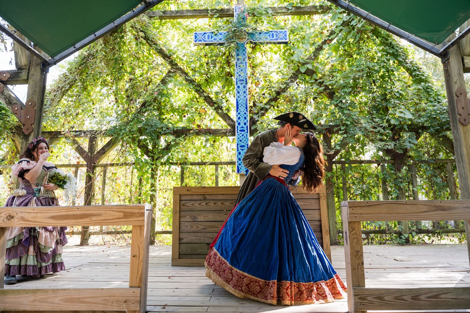 Renaissance wedding ceremony under green canopy with wooden benches and decorative blue pillars.